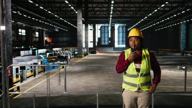 African american engineer holds walkie-talkie and supervising production line in industry plant. Wireless radio device provides strong signal for activity coordination in machinery hall.