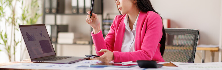 A focused woman in a pink blazer is analyzing data on a laptop, surrounded by documents and tools,...