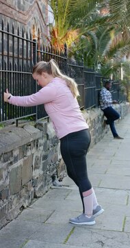 Vertical video: Woman walking to wrought-iron railing on sidewalk and stretching calves for warmup