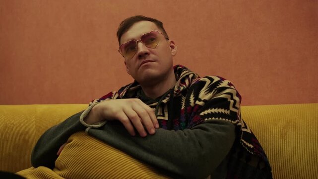Man wearing stylish yellow sunglasses and patterned poncho sitting on yellow sofa. Portrait of relaxed male resting at home in cozy indoor environment. Lifestyle, fashion and leisure concept