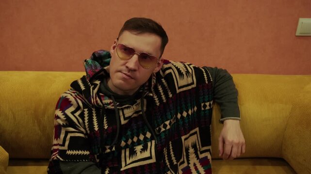 Man wearing stylish yellow sunglasses and patterned poncho sitting on yellow sofa. Portrait of relaxed male resting at home in cozy indoor environment. Lifestyle, fashion and leisure concept