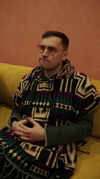 Man wearing stylish yellow sunglasses and patterned poncho sitting on yellow sofa. Portrait of relaxed male resting at home in cozy indoor environment. Lifestyle, fashion and leisure concept