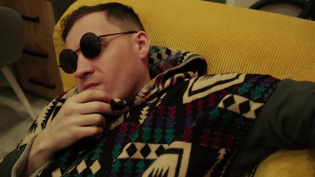 Man wearing ethnic hoodie and round sunglasses resting on couch. Hipster relaxing at home inside yellow interior. Young male in stylish patterned clothing resting on sofa hobby leisure.