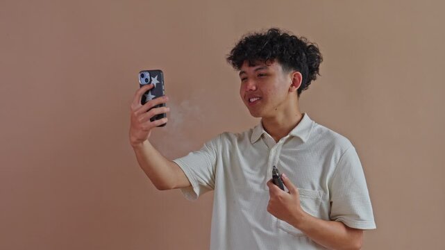 Asian man recording selfie video while vaping with an e-cigarette in studio