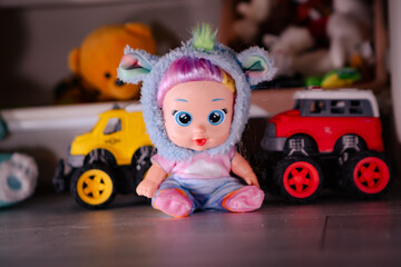Fototapeta premium Little doll wearing blue furry animal hood sitting between toy trucks. Child playing with plastic baby toy on floor. Colorful childhood object collection for gift or nursery decor.