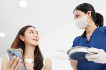Fototapeta premium A woman is looking at her phone while a nurse stands behind her. The nurse is wearing a blue uniform and a mask 