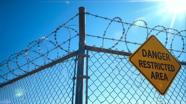 A prominent yellow diamond-shaped warning sign clearly displays "danger restricted area" text, securely affixed to a sturdy chain-link fence, which is heavily fortified with sharp barbed wire coils