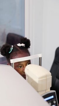 Vertical video, Young African girl child having her vision checked by non-contact tonometer (NCT) in a clinic