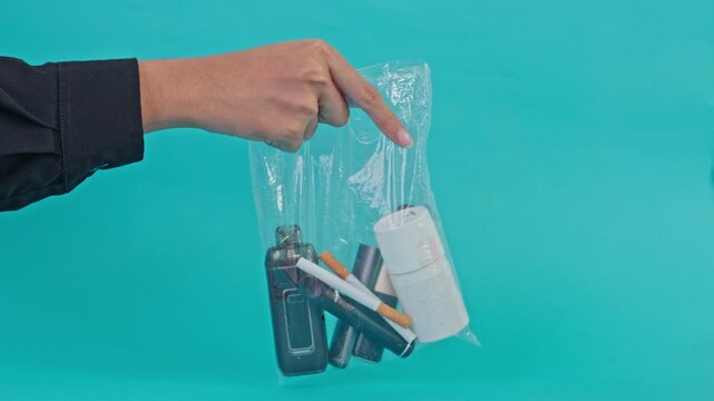 Studio shot of hand carrying plastic bag with various smoking items and white medicine jar on turquoise background
