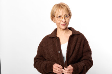 Smiling middle aged woman with short blonde hair and glasses wearing brown coat on white...