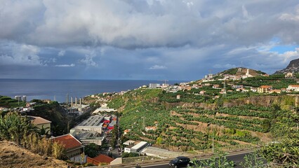 Panoramic coastal landscape of Madeira Island with terraced banana plantations, hillside villages, and the Atlantic Ocean under dramatic clouds, Portugal. © Sebastiaan89