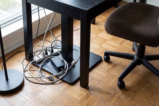A messy tangle of electrical cords and a power strip under a black desk on a wooden floor