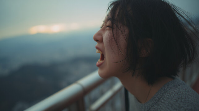 薄暗い夕暮れ時の高台のフェンス際で、遠くの街並みに向かって顔を向け、感情を剥き出しにするように大きく口を開けて叫ぶショートヘアの女性の横顔のアップ