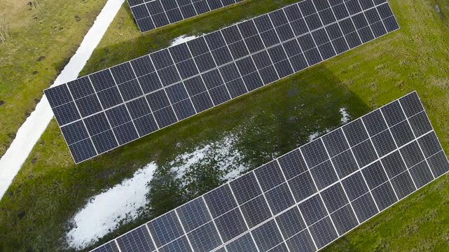 Drone orbit above solar panel rows generating renewable electricity on spring grass with visible melting snow patches.