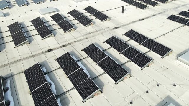 Low altitude drone view orbiting large rooftop solar panel arrays with visible wiring and small snow patches on a bright spring day.