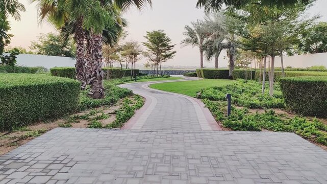 Walking path with lush green landscaping, featuring carpet grass, trees, palms and Washingtonias. Villas houses alongside greenery garden backyard. Interlocking stone pathway weaves through landscape.