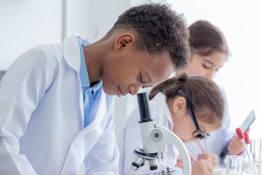 Close-up group of multicultural cute elementary students kids wearing lab coat uniforms using microoscope writing experiment report in laboratory room, lifestyle pupil boy and girls learning education