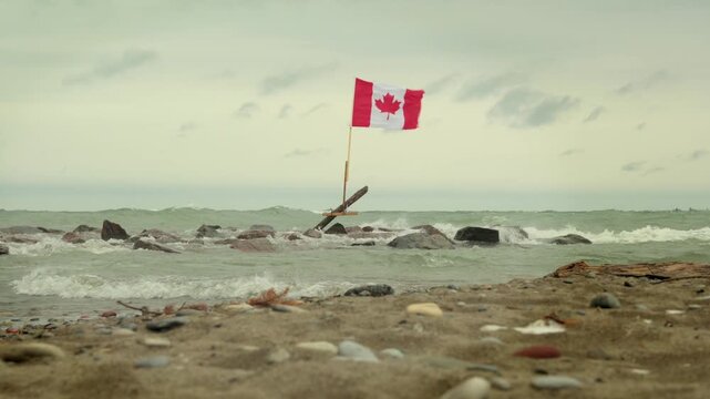 A Canadian flag stands on a rocky Lake Ontario shoreline on an overcast winter day, with waves washing over the beach in Toronto, Ontario, Canada.