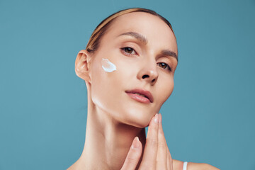 Woman applying white face cream on cheek with fingers. Close-up portrait on blue background....