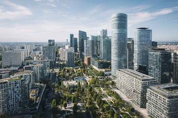 Obraz premium Aerial view of modern city skyline with tall office buildings and green park area under clear blue sky, showcasing urban architecture and vibrant city life