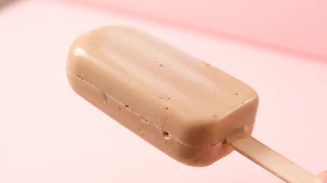 Close-up of gourmet creamy coffee or caramel ice cream popsicle bar on a wooden stick, highlighting the smooth texture against a vibrant pink background.