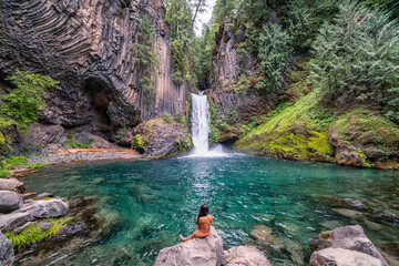 A woman is sitting in a body of water, looking out at a waterfall © ExploringandLiving
