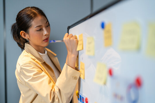 Asian female UX/UI designer writing on sticky notes on whiteboard, Young creative professional brainstorming user journey, empathy mapping and digital product design bootcamp concept