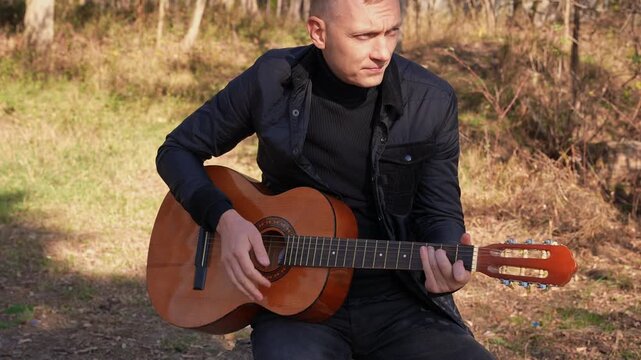 Beautiful blonde man playing acoustic guitar and singing relaxing enjoying outdoors on sunny autumn day. Artist learning and practicing playing musical instrument. Fall leaves autumnal weather 
