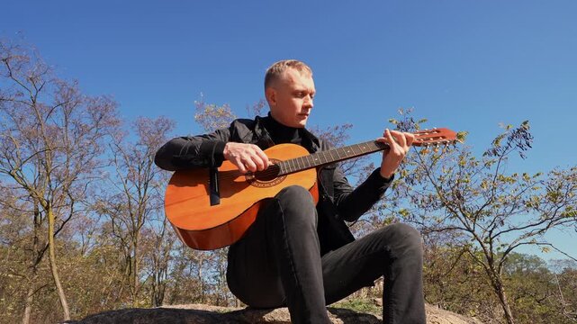 Blonde man playing string guitar outdoors in autumn forest. Concept of sound therapy, mental health and wellness rituals. Calmness tranquility audio-sensory practices. Aura farming energy holistic