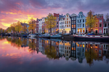 Fototapeta premium Amsterdam canal panorama with Dutch houses and boats at sunset reflects colorful sky and autumn trees, creating peaceful and picturesque urban scene by water