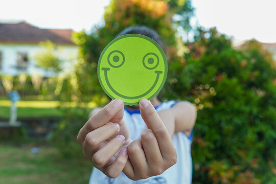 Person holding a green smiley face paper circle covering their face in a sunny garden