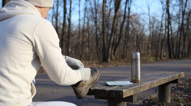 Person in a hoodie sitting on a wooden bench, tying hiking boot laces with a map and thermos nearby in a park.