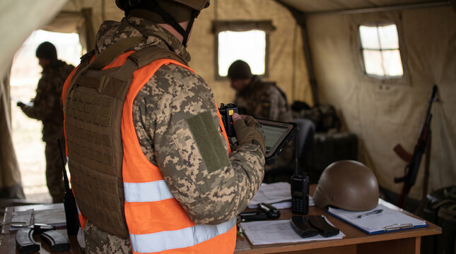 Military officer in camouflage uniform and orange vest using a digital tablet inside a tactical command tent.