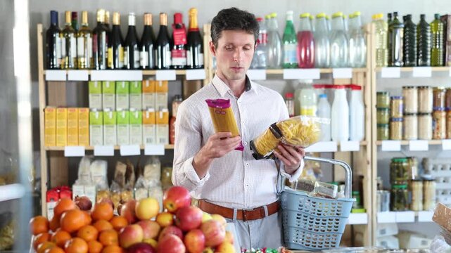 Man in light shirt took some varieties of pasta from shelf. Client compare pasta noodles items package. Customer examines contents of package, choose noodles garnish product