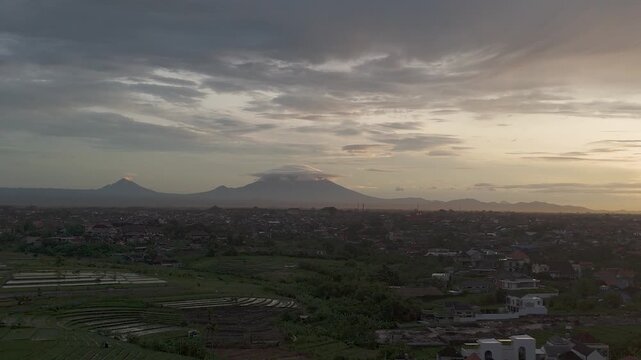 Cinematic aerial view of Mount Agung and Mount Abang at sunrise, overlooking lush rice fields and residential areas in Bali, Indonesia. 4K footage.