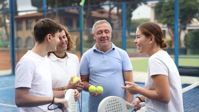 Happy laughing men and woman of different ages in sportswear with rackets for padel and balls in hands talking friendly near net 