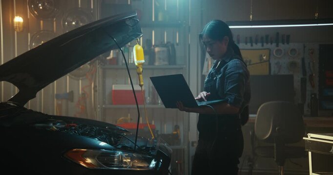 Woman Automotive Technician Stands Open Car Hood, Holding Laptop Connected Vehicle Onboard Computer System. Girl Methodically Scanning For Engine Faults and Monitoring Real Time Data on Screen.