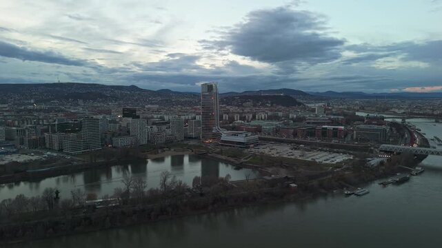 Orbital aerial view of Budapest at sunset, highlighting the Mol Tower, Danube River, surrounding urban architecture, and scenic cityscape with reflections, traffic, and the Buda Hills in the distance.