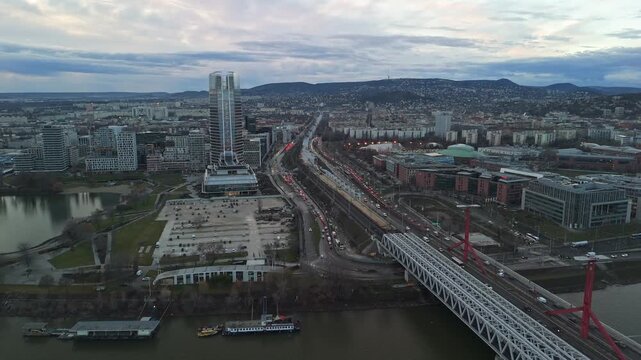Backward drone view of Budapest at sunset, highlighting the Mol Tower, Danube River, L&aacute;gym&aacute;nyosi Bridge, and surrounding urban architecture, with traffic and cityscape blending into a scenic panorama.