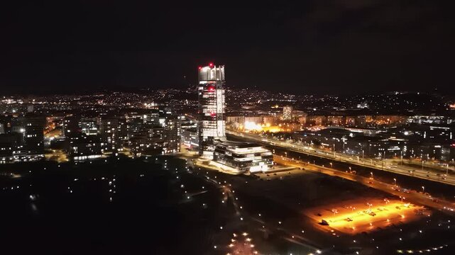 Aerial nighttime view of Budapest featuring the illuminated Mol Tower, vibrant city lights, surrounding urban landscape, and visible traffic in Hungary's bustling capital.