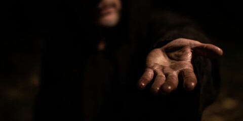 Religious figure reaching out hand in dark studio setting for spiritual church advertising, hooded robed man offering help with copy space for inspirational content