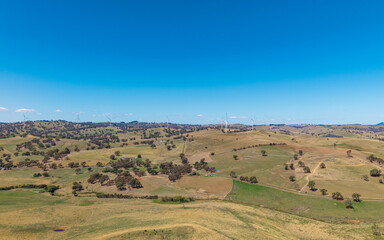Fototapeta premium Expansive Aerial View of Wind Turbines on Verdant and Golden Rolling Hills Under a Clear Blue Sky