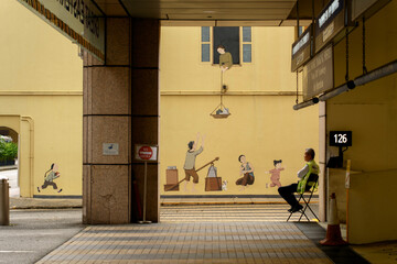 Security guard resting near a colorful mural depicting traditional life in Chinatown district. Street art and everyday urban scene in Singapore. © dron285