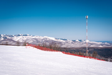 A snowy ski slope with a red safety fence overlooks distant mountain ranges under clear sky
