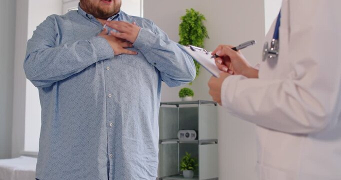 Overweight patient holds his chest, describing sharp pain to cardiologist doctor at medical clinic, noting heartburn. Fat obese man reports chest pain during healthcare consultation.