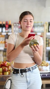 Woman in store scans barcode QR code on tin cans of preserves. With help of mobile app, buyer receives information about product. She checks manufacturers data in registry database of national agency