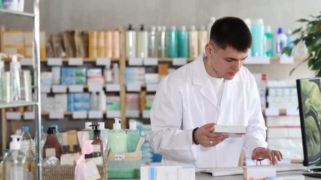 Near counter with computer is pharmacists workplace. Guy employee participates in online testing and learning, reads incoming emails. Electronic database of drugs. Colleague work in background