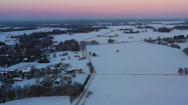 Winter expanse covering houses, fields, and roads during twilight glow in aerial view with snowfall and pink sky