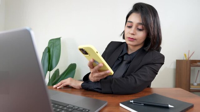 Close up of beautiful young Indian business woman sits on chair at workplace desk using smart phone business communication, checking online email messages sms.