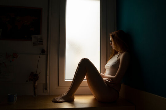Serious young woman sitting on windowsill at home